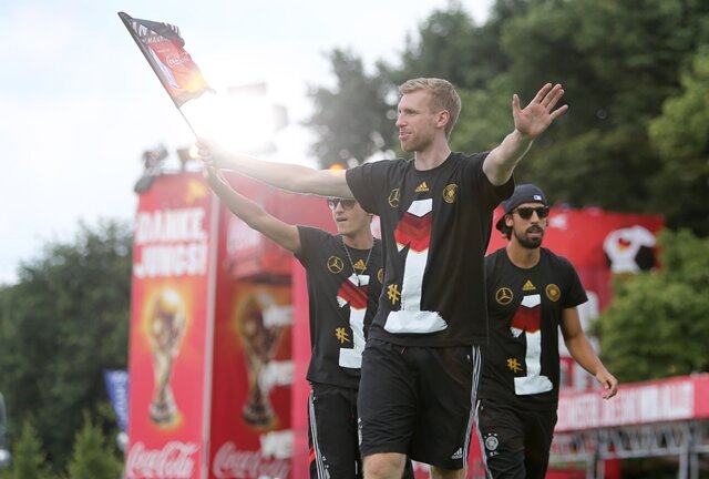 <p>
	Per Mertesacker feiert auf der Bühne mit der Flagge in der Hand. Mesut Özil und Sami Khedira stehen links und rechts von ihm.</p>
