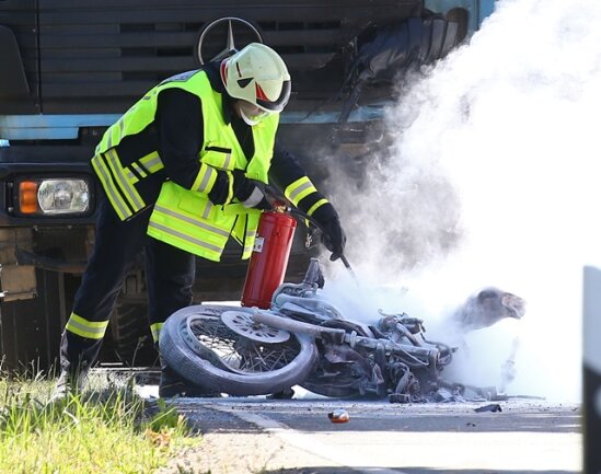 <p>
	Ein Feuerwehrmann löscht das brennende Motorrad, das vor einem Brummi liegt. Dichte Qualmwolken ziehen über die Straße.</p>
