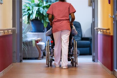 Eine Bewohnerin eines Pflegeheims, die im Rollstuhl sitzt, wird von einer Pflegerin den Gang entlang geschoben.