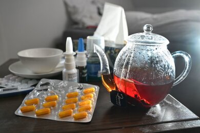 Medikamente und eine Kanne und Tasse Tee zur Bekämpfung einer Erkältung stehen auf einem Nachttisch.
