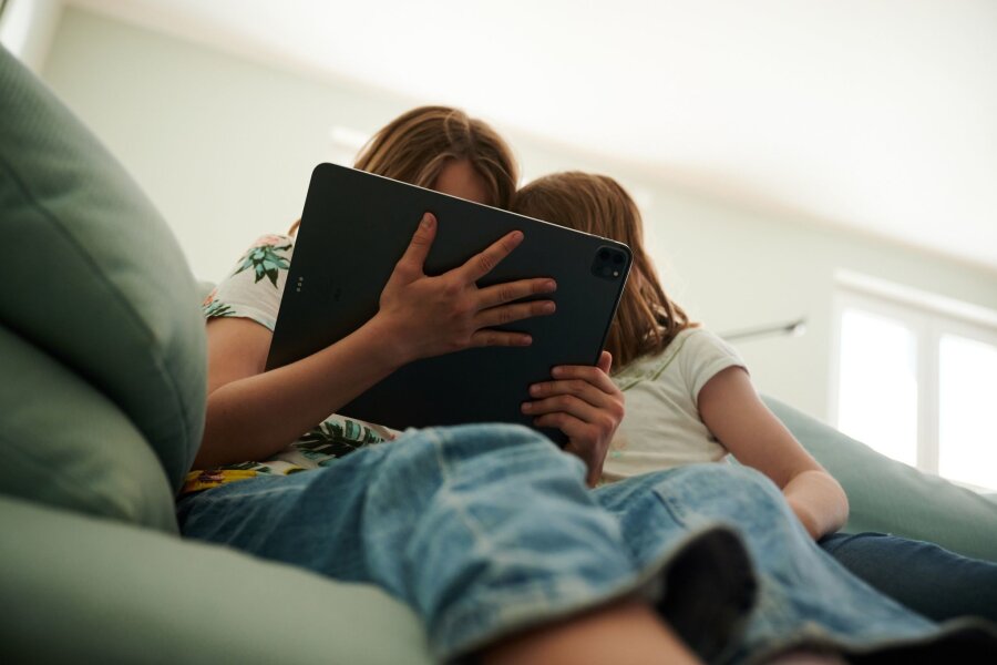 73 Prozent der Kinder unter 18 Jahren leben mit Geschwistern - Zwei Schwestern sitzen auf der Couch und gucken in das Tablet ihrer Eltern.