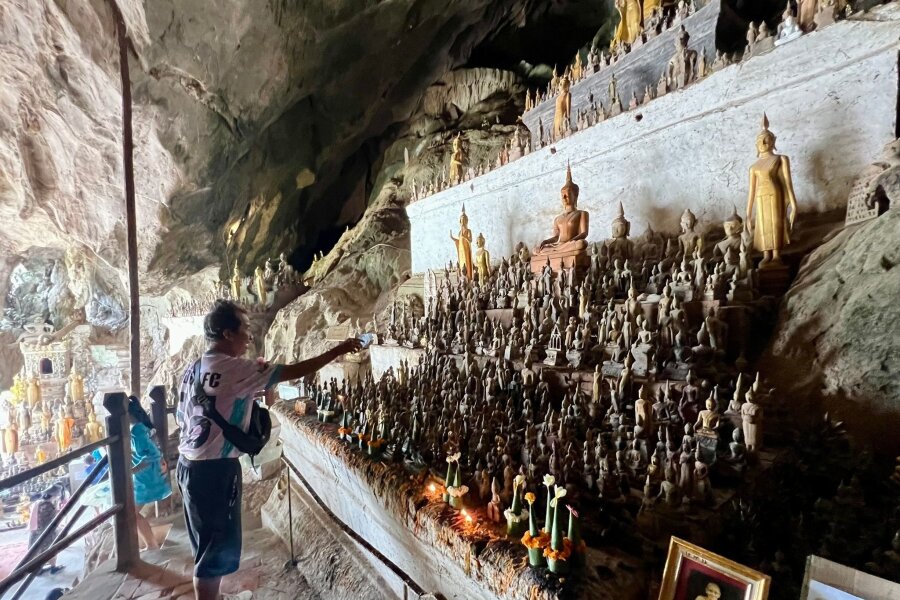 Am Mekong: Heilige Höhlen mit 6000 Buddha-Statuen - Ein Mann besprenkelt Buddha-Statuen in den Pak Ou Caves als Zeichen des tiefen Respekts mit Wasser.