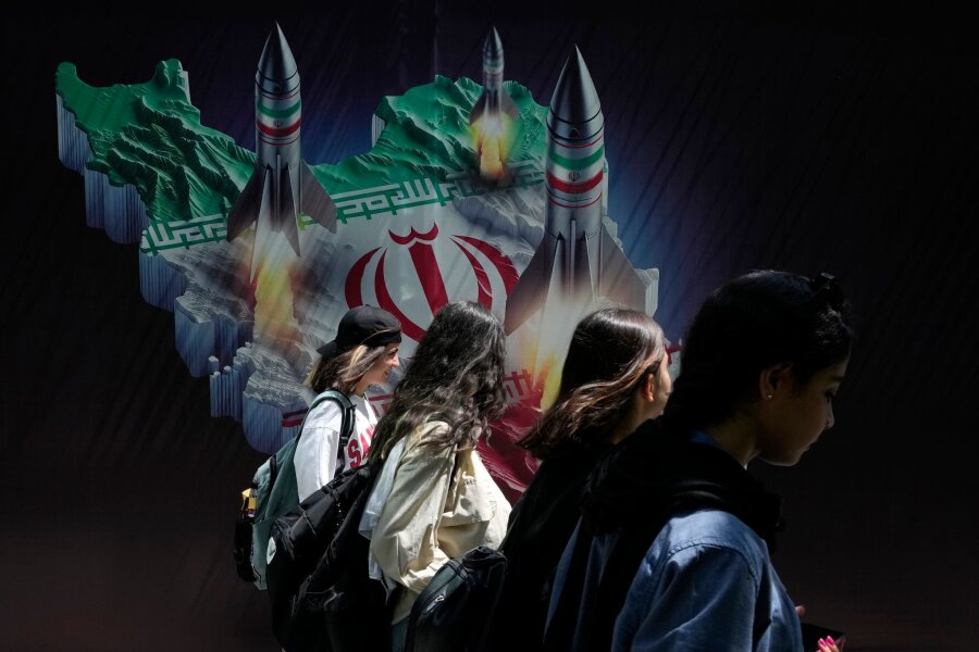 Bilder des Tages vom 19.04.2024 - Iranische junge Frauen, die ihr obligatorisches islamisches Kopftuch nicht tragen, gehen in Teheran an einem Banner vorbei, das den Abschuss von Raketen auf einer iranischen Landkarte zeigt.