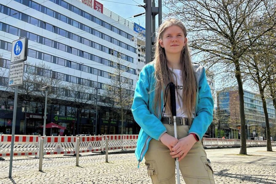 Im Hintergrund sieht man die Baustelle am Roten Turm in Chemnitz. Davor steht eine junge Frau mit Blindenstock. Sie trägt eine hellbraune Hose ein weißes Shirt und eine türkisfarbene Jacke.