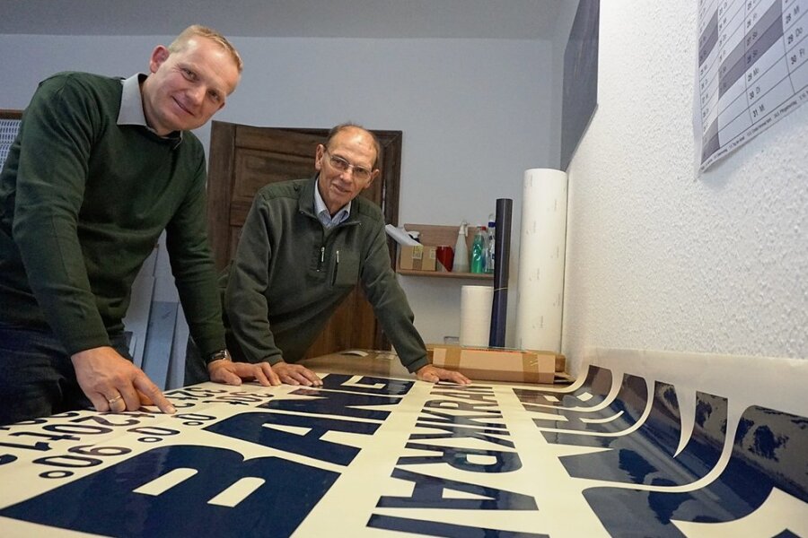 Vor 30 Jahren gründete Wolfgang Röh (rechts) den Werbemarkt Röh in Bad Elster. Sein Sohn Thomas Röh stieg 2011 in das Geschäft ein und hat es im Jahr 2014 von seinem Vater übernommen. Das Bild zeigt beide an einem aktuell produzierten Aufkleber für die Oelsnitzer Firma Bang Kransysteme GmbH.