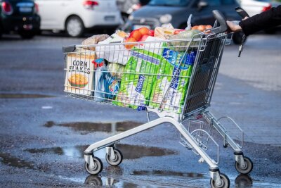 Ein Kunde schiebt einen bis zum Rand gefüllten Einkaufswagen über den Parkplatz eines Supermarktes.