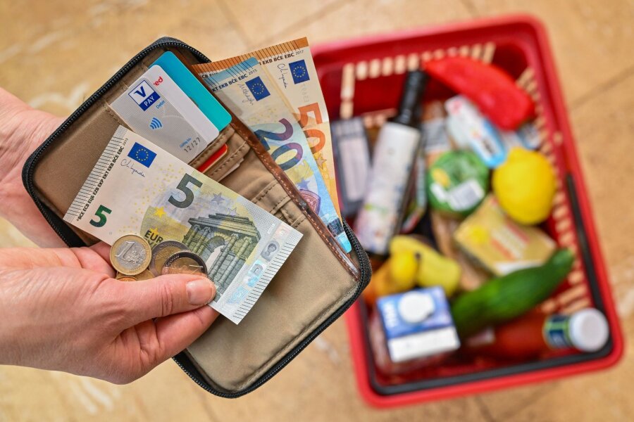Inflationsrate in Sachsen über 3 Prozent - Eine Frau hält auf ihrer Hand Geld vor einem vollen Einkaufskorb mit Lebensmitteln.