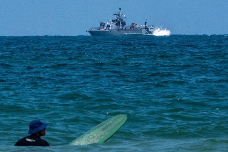 Iran und Israel nach Direktschlag am Rande des Abgrunds - Ein Surfer wartet auf eine Welle, während ein israelisches Marineschiff im Mittelmeer vor der Küste von Hadera patrouilliert.