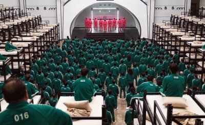 Netflix-Serie "Squid Game" weckt in Zwickau Interesse an Korea - Die Antagonisten in "Squid Game": Die Teilnehmer des Spiels in dunkelgrünen nummerierten Trainingsanzügen, die Wächter in rote Kapuzen-Overalls und schwarzen Fechtmasken.