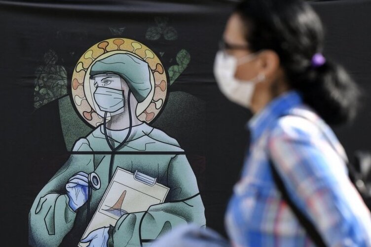 Eine Frau mit Schutzmaske läuft an einem Straßenplakat vorbei, das medizinisches Personal in Gestalt einer Heiligen darstellt