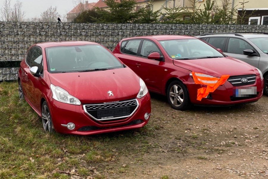 Seit einem Jahr steht auf dem Parkplatz ein roter Peugeot ohne Kennzeichen, daneben ein roter Kia, bei dem eine Warnweste unter der Motorhaube klemmt. 