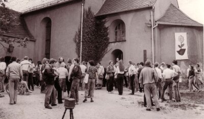 Themen ändern sich - Idee bleibt - Teilnehmer des Friedensseminars in den 80er-Jahren vor der Kirche Königswalde, rechts an der Fassade das Symbol des Treffens, der umgedrehte DDR-Stahlhelm aus dem eine Sonnenblume wächst.