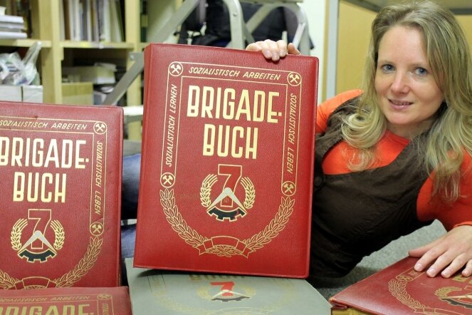 
              <p class="artikelinhalt">Mitarbeiterin Ina Georgi präsentiert die alten Brigadebücher der Wismut mit dem Symbol des Sieben-Jahres-Planes in der DDR. Sie werden ausgewertet und in die Bibliothek aus mehr als 2000 Büchern eingeordnet, die im Moment im Entstehen ist. </p>
            